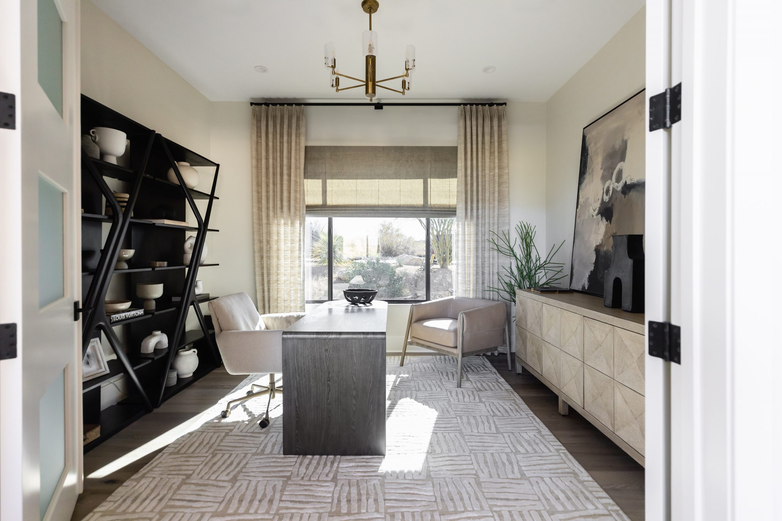 A home office with a modern desk, two chairs, shelving, a large sideboard, abstract art, and a curtained window.— Living with Lolo.