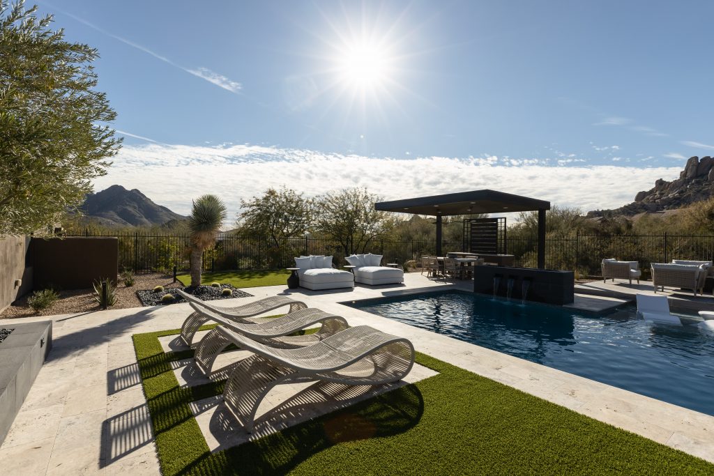 Modern outdoor pool area with lounge chairs, sunbeds, a covered bar, and mountain views under a clear sky—Living with Lolo.