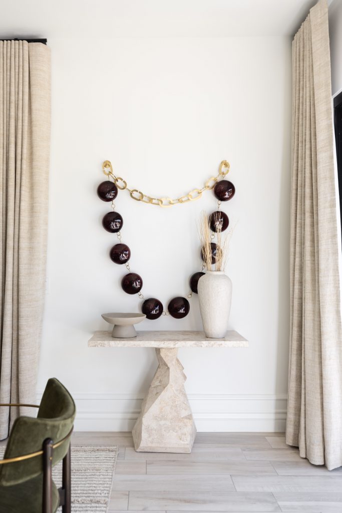 A chain with dark links and gold connectors hangs on a white wall above a stone console table with a vase and bowl. Beige curtains frame the scene.— Living with Lolo.
