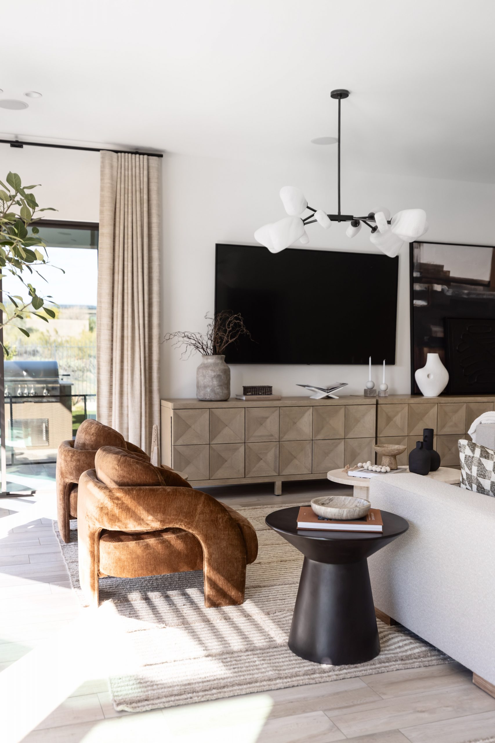 Modern living room with a wall-mounted TV, wooden cabinet, two brown chairs, black side table, and large windows with beige curtains—Living with Lolo.