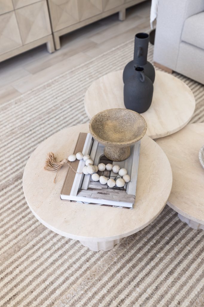 A round beige coffee table holds stacked books, a wooden bead garland, a bowl, and two black vases on a striped rug—Living with Lolo.