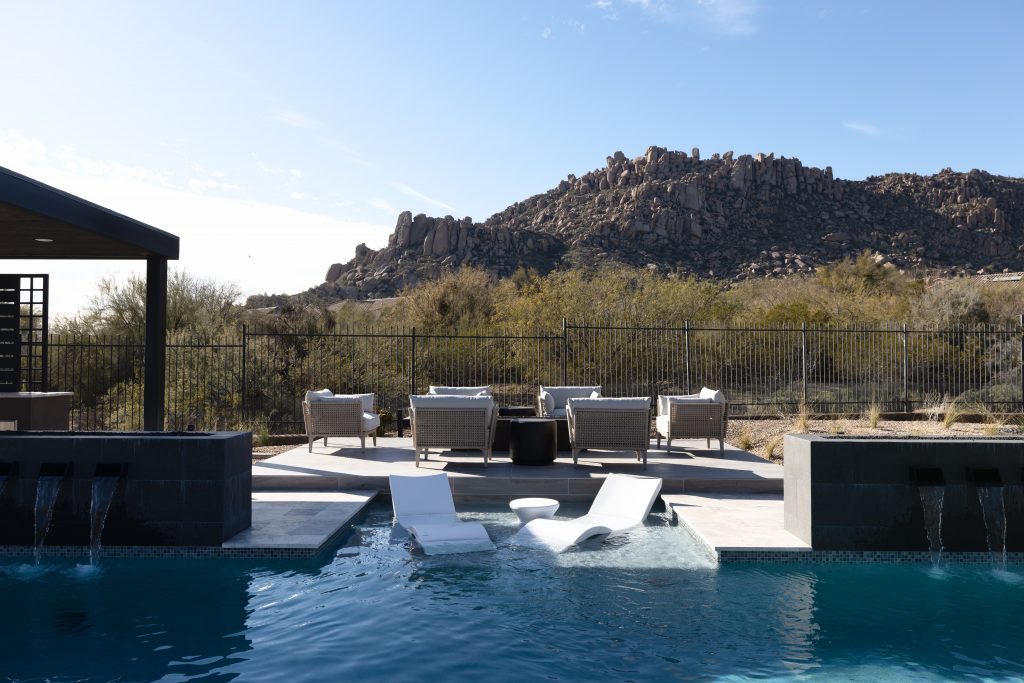 A modern pool with two white lounge chairs in the water and a patio seating area adjacent to the home—Living with Lolo.