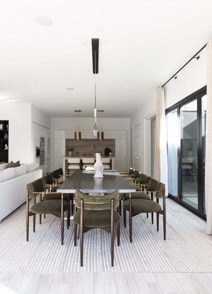 Modern dining room with a long black table, eight green velvet chairs, a white vase centerpiece, light rug, and large glass doors—Living with Lolo.