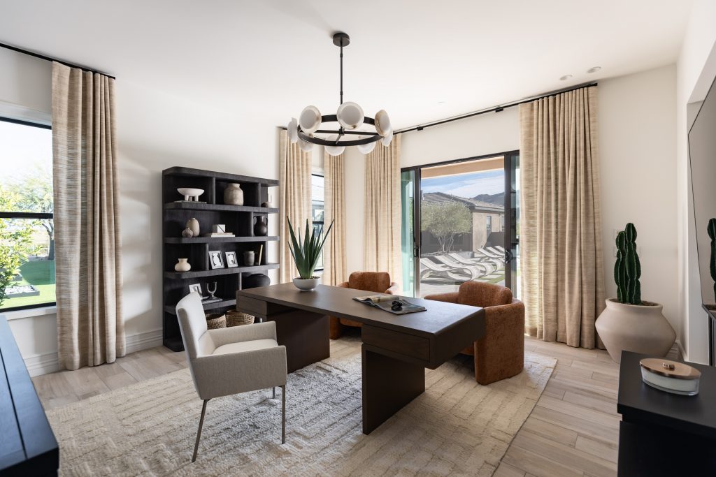 Modern home office featuring a large desk, white chair, brown armchairs, black bookshelf, and large windows with beige curtains—Living with Lolo.