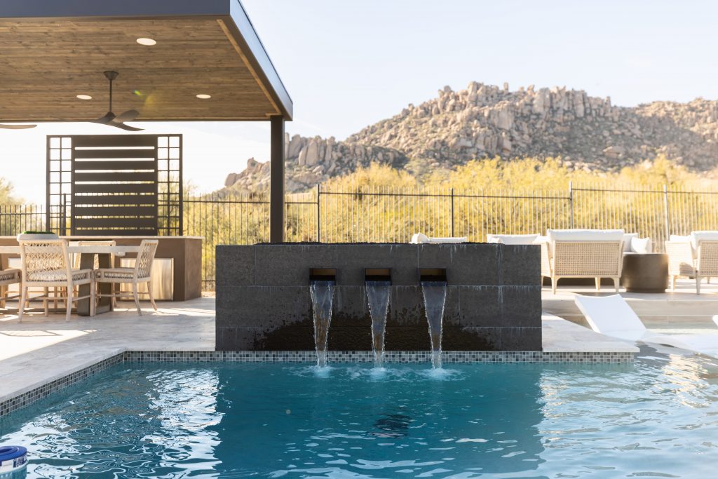 Modern outdoor pool featuring a black stone water element, lounge chairs, shaded seating area, and rocky landscape beyond— Living with Lolo.