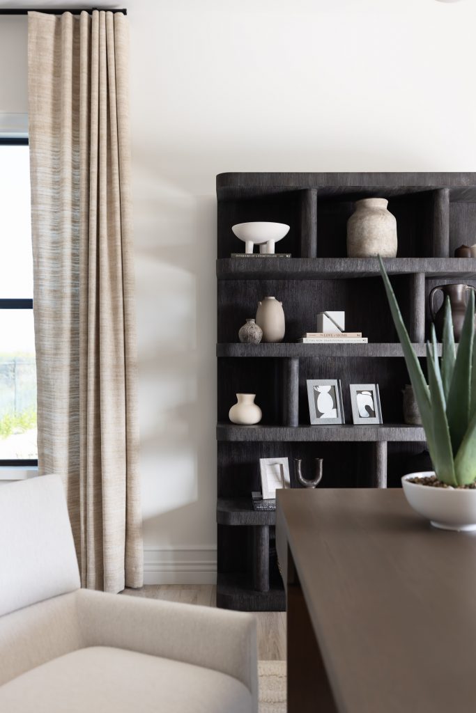 A beige armchair sits beside tall light curtains, a dark wooden bookshelf with decor, and a potted plant on a dark table. — Living with Lolo.