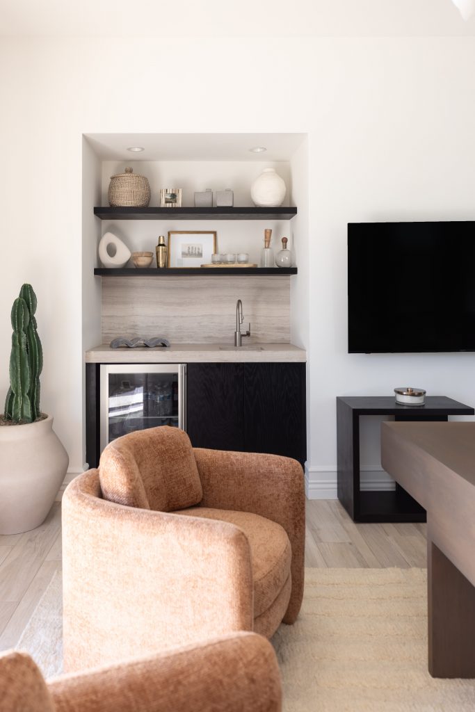Built-in wet bar with open shelves, a small sink, mini fridge, brown swivel chair, potted cactus, and wall-mounted TV—Living with Lolo.