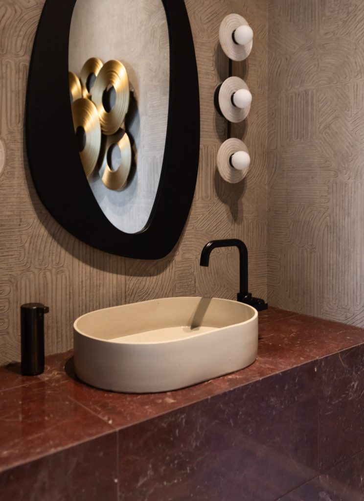 A bathroom with a black faucet, oval mirror, three wall lights, gold accents, and a dark red marble countertop.— Living with Lolo.