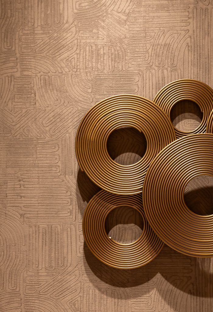 Gold circular metal wall art with concentric rings is mounted on a textured, patterned beige wall.— Living with Lolo.