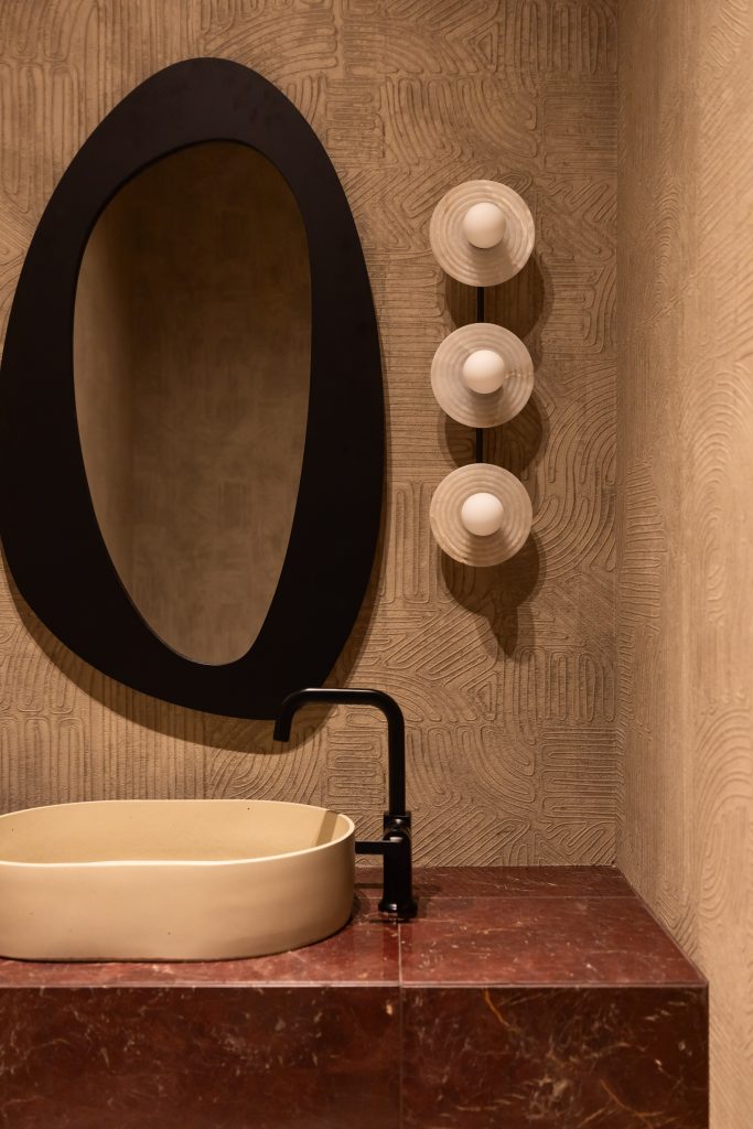 An oval mirror hangs above a beige vessel sink with a black faucet on a red marble countertop, lit by three round wall lights—Living with Lolo.