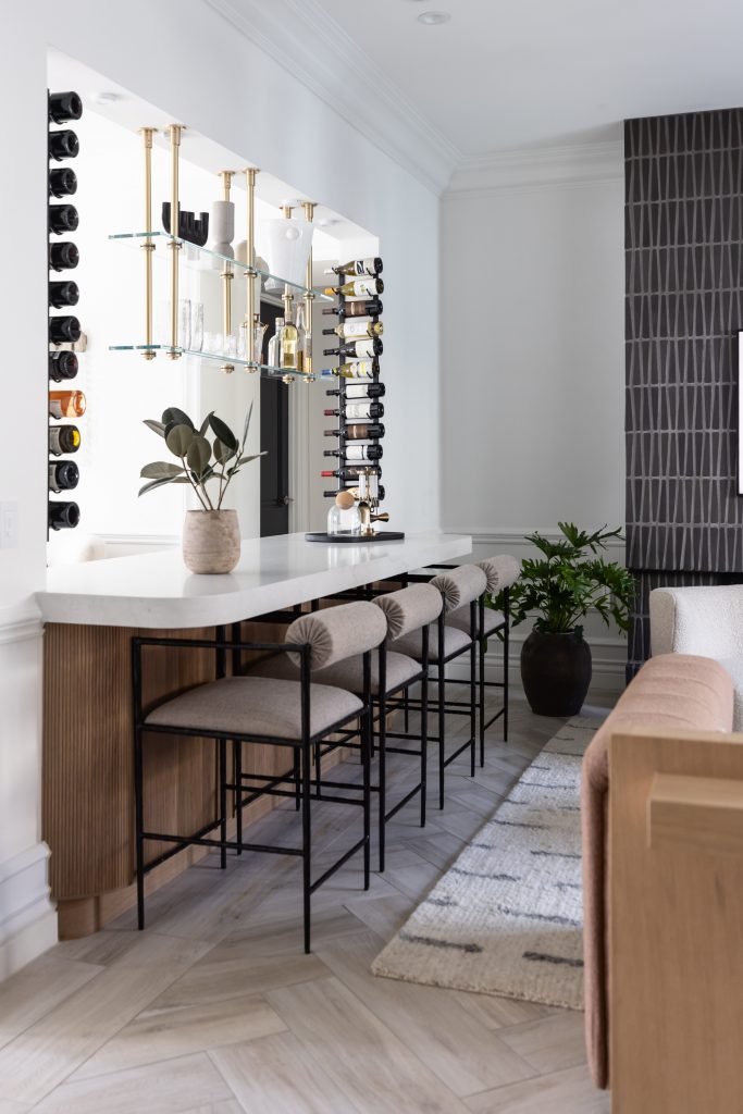 Modern home bar area featuring a white countertop, three upholstered bar stools, a potted plant, and wall-mounted glass shelves—Living with Lolo.