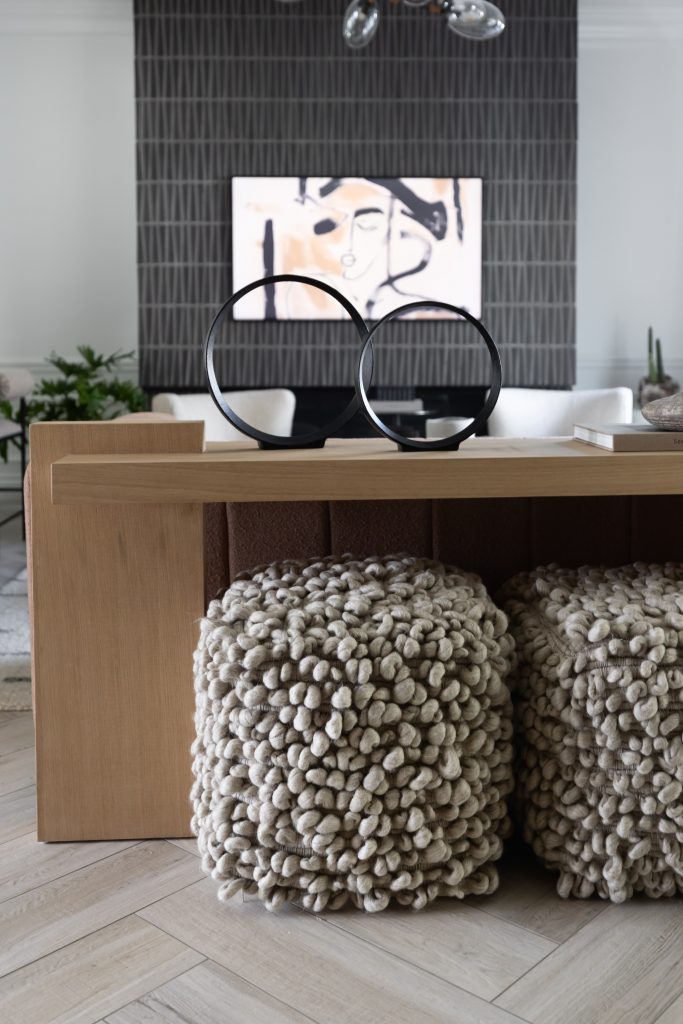 A modern living area with a wooden console table, two chunky knit poufs, circular black sculptures, and abstract art on a tiled accent wall. — Living with Lolo.