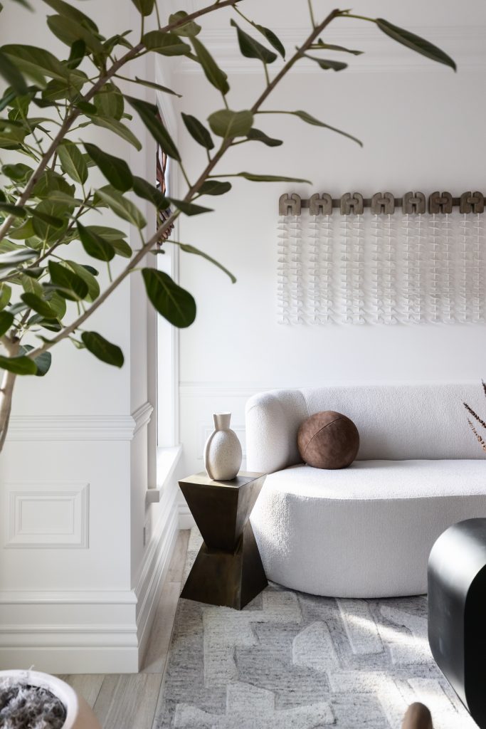 A modern living room with a white curved sofa, round brown cushion, geometric side table with a vase, large plant, and textured wall art—Living with Lolo.