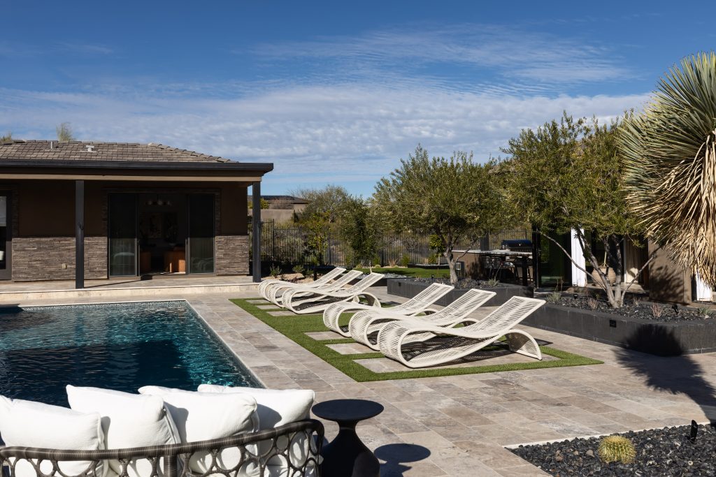Outdoor pool area featuring curved lounge chairs on artificial grass, tiled decking, patio furniture, and landscaped trees—Living with Lolo.