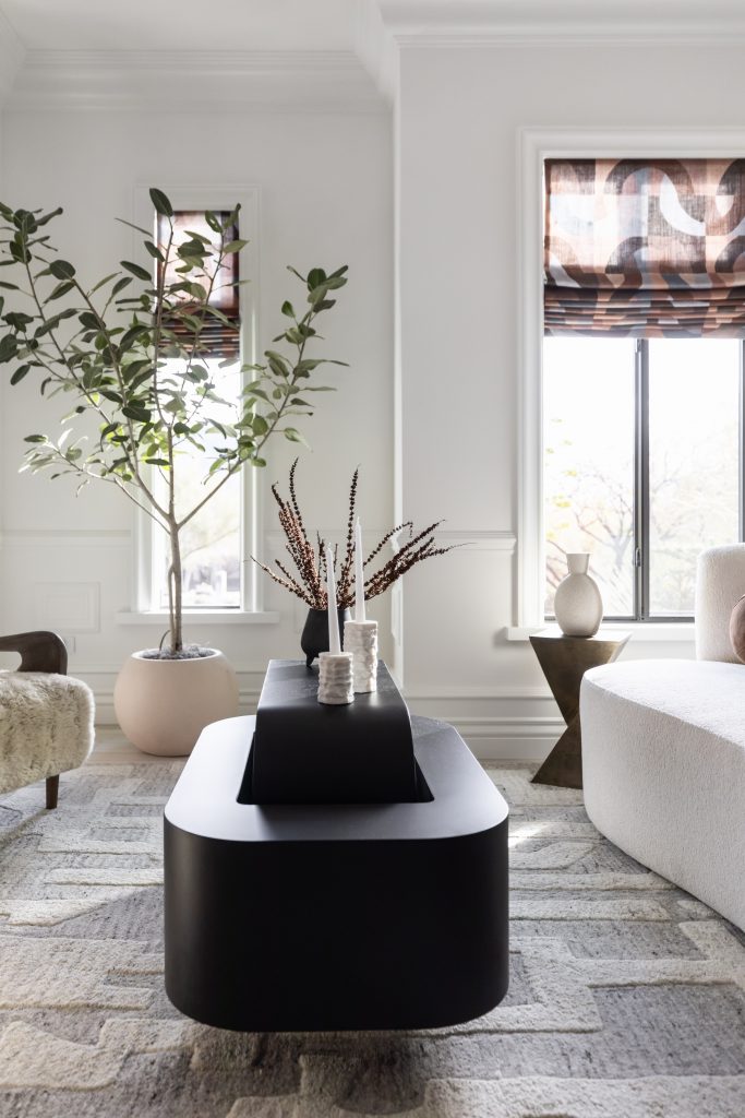 A black coffee table, decorative vases, potted plant, textured rug, white sofa, and two windows with patterned blinds. — Living with Lolo.