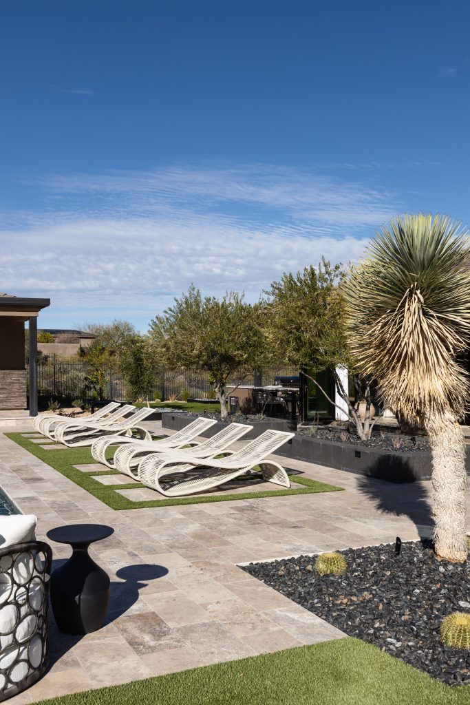 A stone patio with four modern lounge chairs sits next to a pool, surrounded by desert plants and cacti.— Living with Lolo.