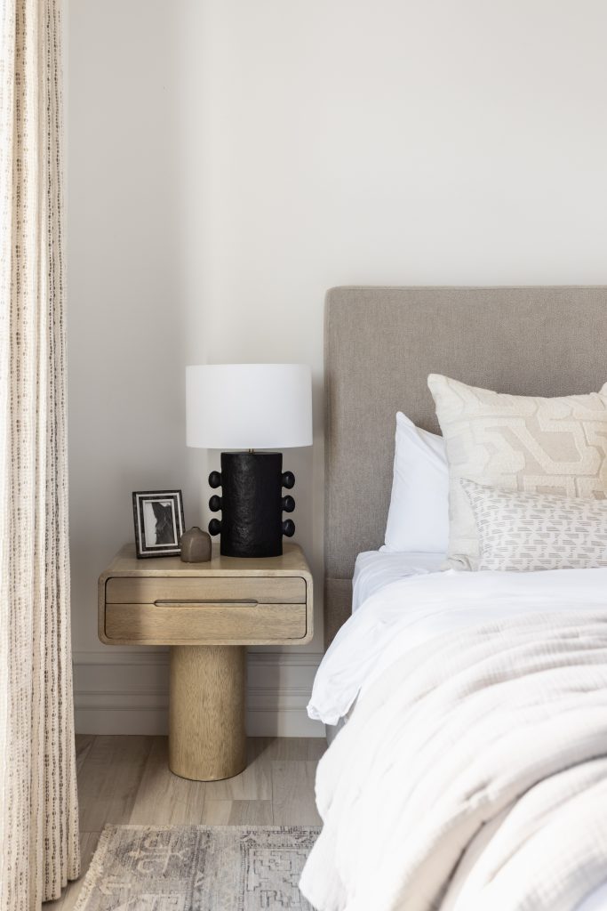 A modern bedside table with a lamp, framed photo, and decorative object is next to a bed with neutral bedding—Living with Lolo.