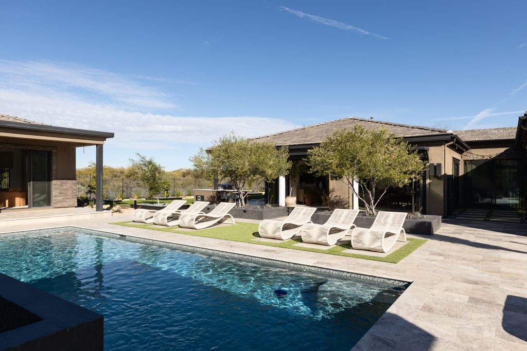 A modern outdoor pool area features six lounge chairs on artificial grass, bordered by contemporary homes and trees. — Living with Lolo.
