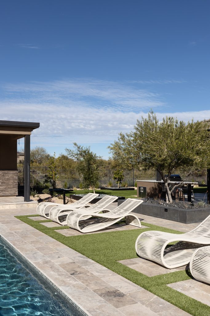 Four modern white lounge chairs rest on artificial grass next to a stone-tiled pool, with trees and a barbecue area visible—Living with Lolo.