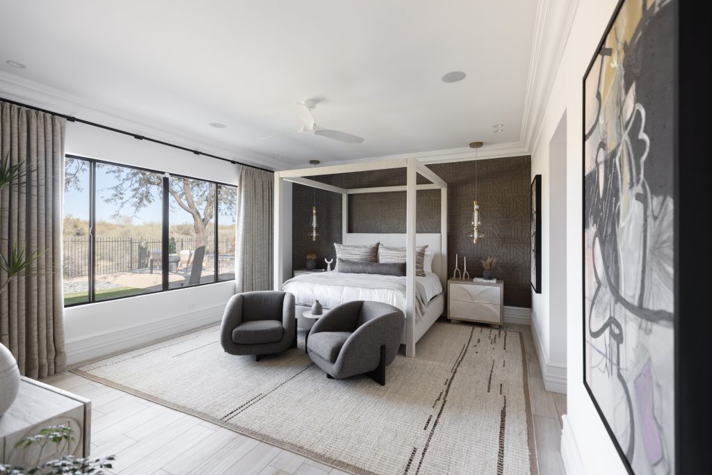 Modern bedroom featuring a four-poster bed, two gray armchairs, large curtained windows, neutral decor, and abstract wall art—Living with Lolo.