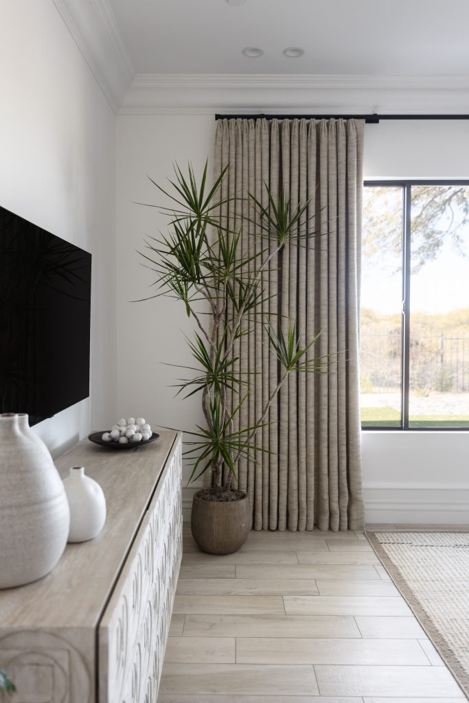 A potted dracaena plant sits by a window with beige curtains, next to a light wood console and decorative vases—Living with Lolo.