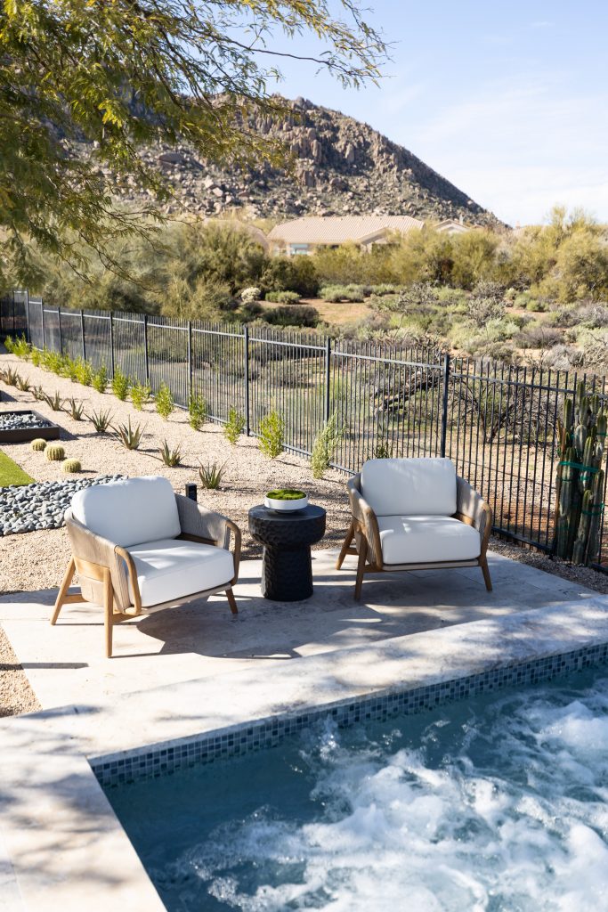 Two cushioned patio chairs and a small side table are arranged next to a pool, bordered by a black metal fence.— Living with Lolo.