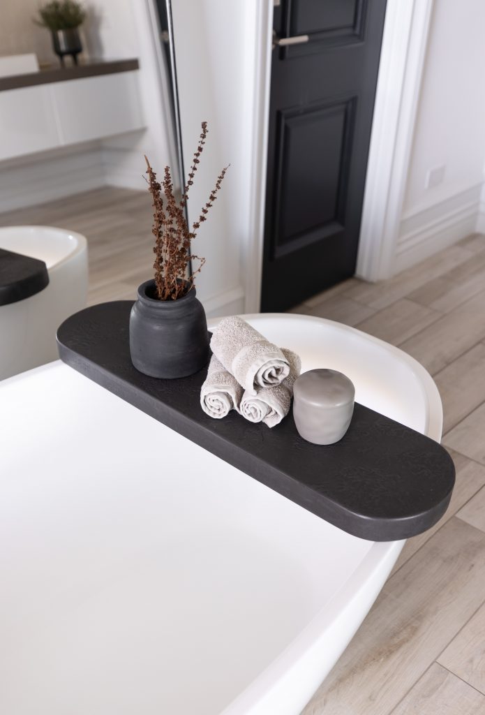 A black bathtub tray on a white tub displays a vase with dried branches, three beige towels, and a gray container—Living with Lolo.