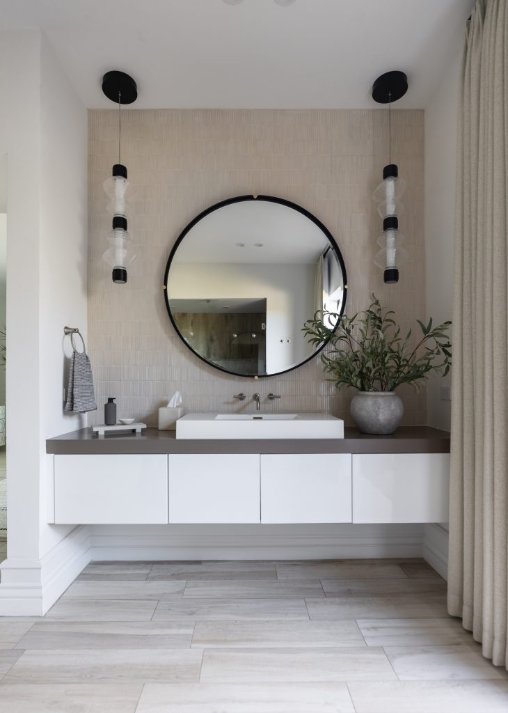 A bathroom with a floating white vanity, round black-framed mirror, pendant lights, potted plant, beige tile wall, and wood floor. — Living with Lolo.