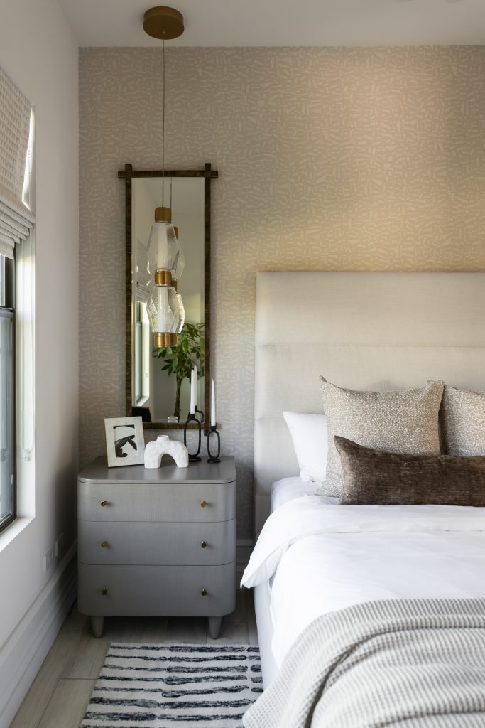 A neatly made bed with neutral bedding is next to a gray nightstand holding decor, candles, and framed art. A mirror and hanging light are above, with a window on the left—Living with Lolo.