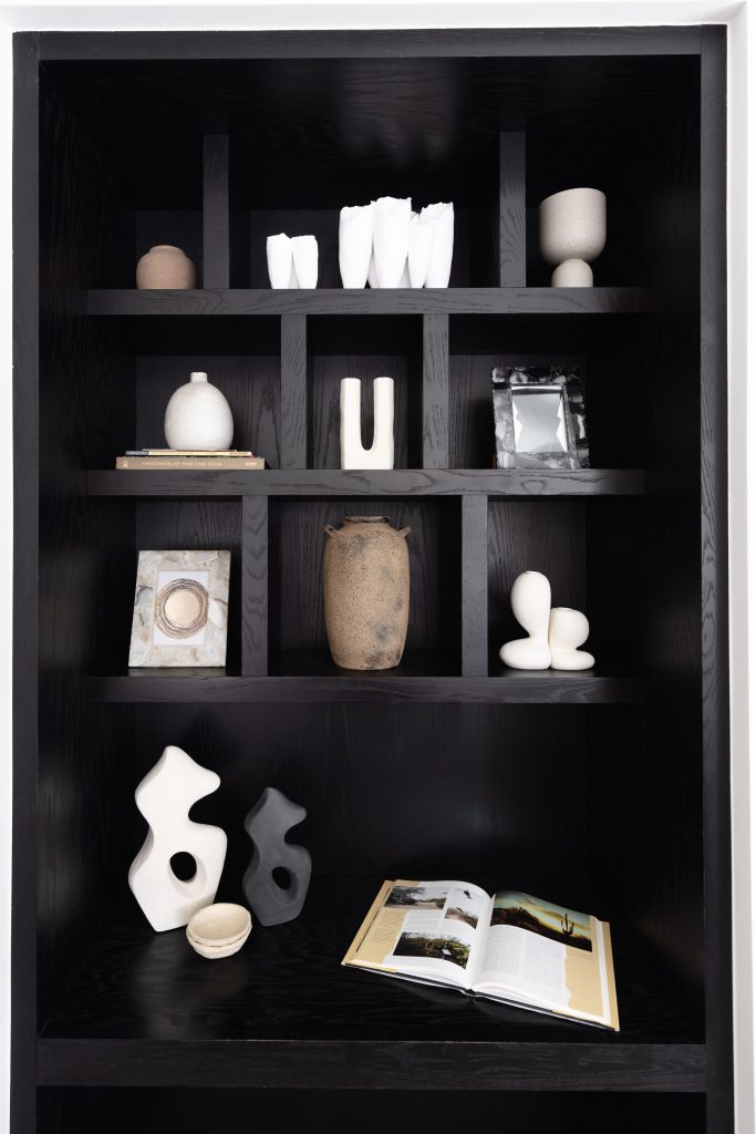 A black shelving unit holds ceramic sculptures, vases, framed art, and an open book with photos and text. — Living with Lolo.
