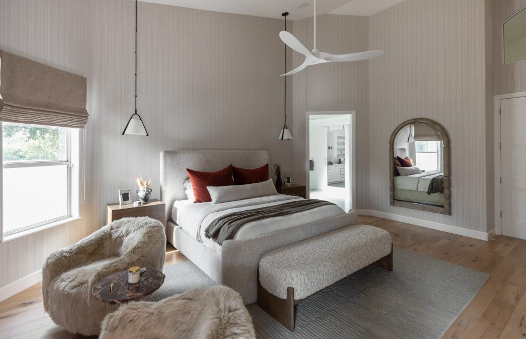 Modern bedroom featuring a large bed, bench, side table, plush chairs, pendant lights, ceiling fan, window shade, and wall mirror—Living with Lolo.
