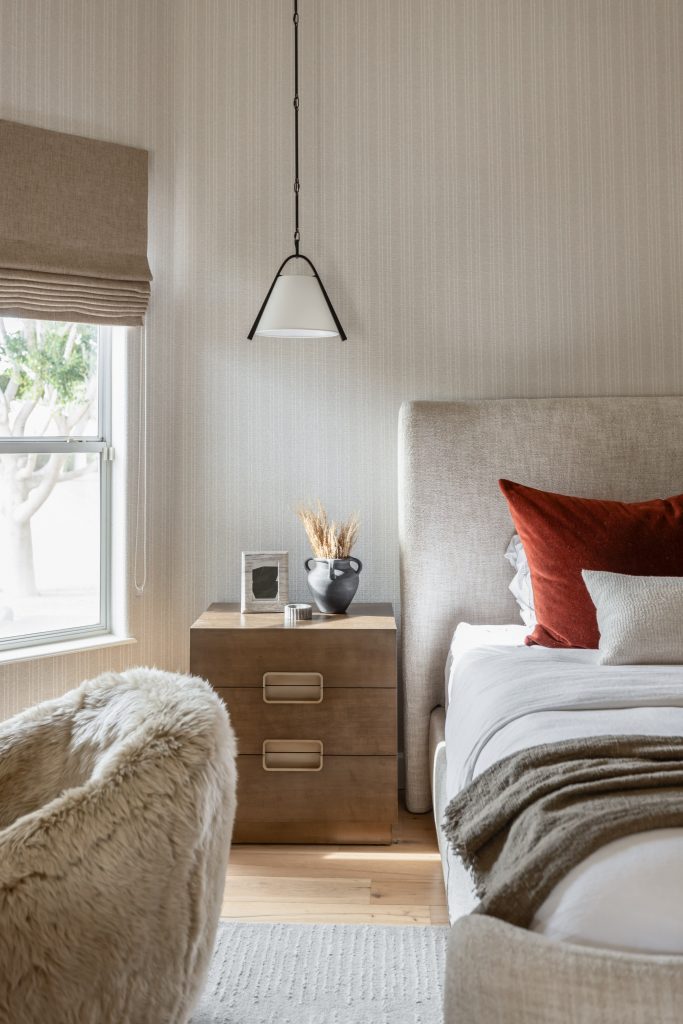 A modern bedroom with a beige upholstered bed, brown and white bedding, a wooden nightstand, fur chair, and a window with a Roman shade. — Living with Lolo.