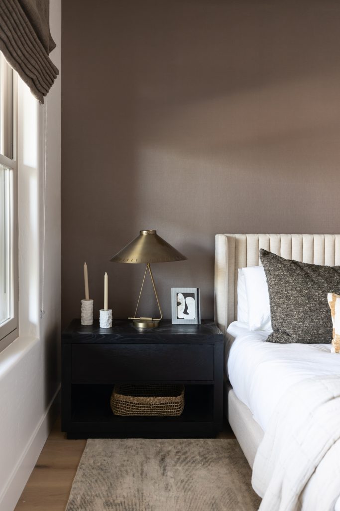 A modern nightstand holds two candles, a framed picture, a brass lamp, and a woven basket on the lower shelf next to a bed—Living with Lolo.