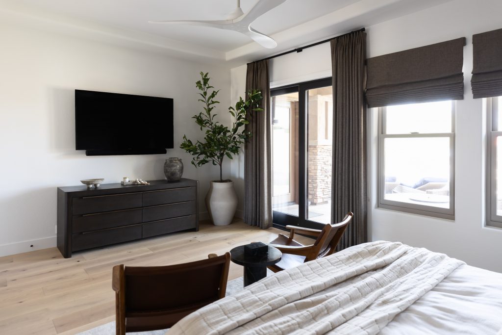 A bed, two wooden chairs, a dresser with a TV above, large potted plant, and windows with brown curtains are visible—Living with Lolo.