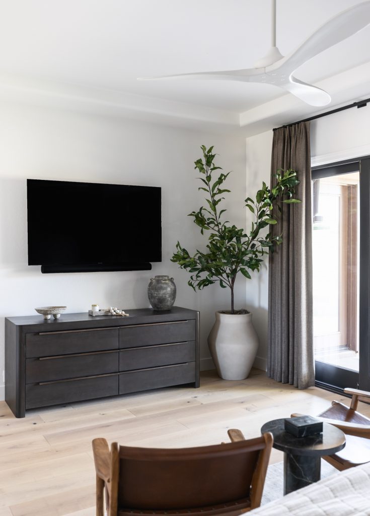 A wall-mounted TV, dark dresser, large potted plant, and floor-to-ceiling curtains by glass doors are shown—Living with Lolo.