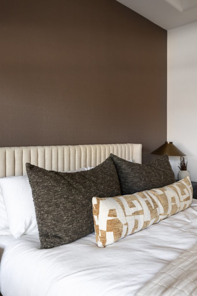 A neatly made bed with a beige upholstered headboard, white bedding, two brown pillows, a patterned bolster, and a lamp on a nightstand—Living with Lolo.