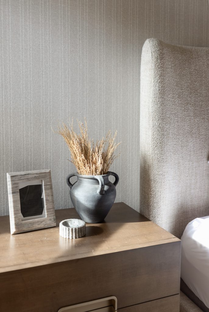A wooden nightstand holds a black vase with dried grass, a small round ornament, and an empty picture frame beside a beige bed—Living with Lolo.
