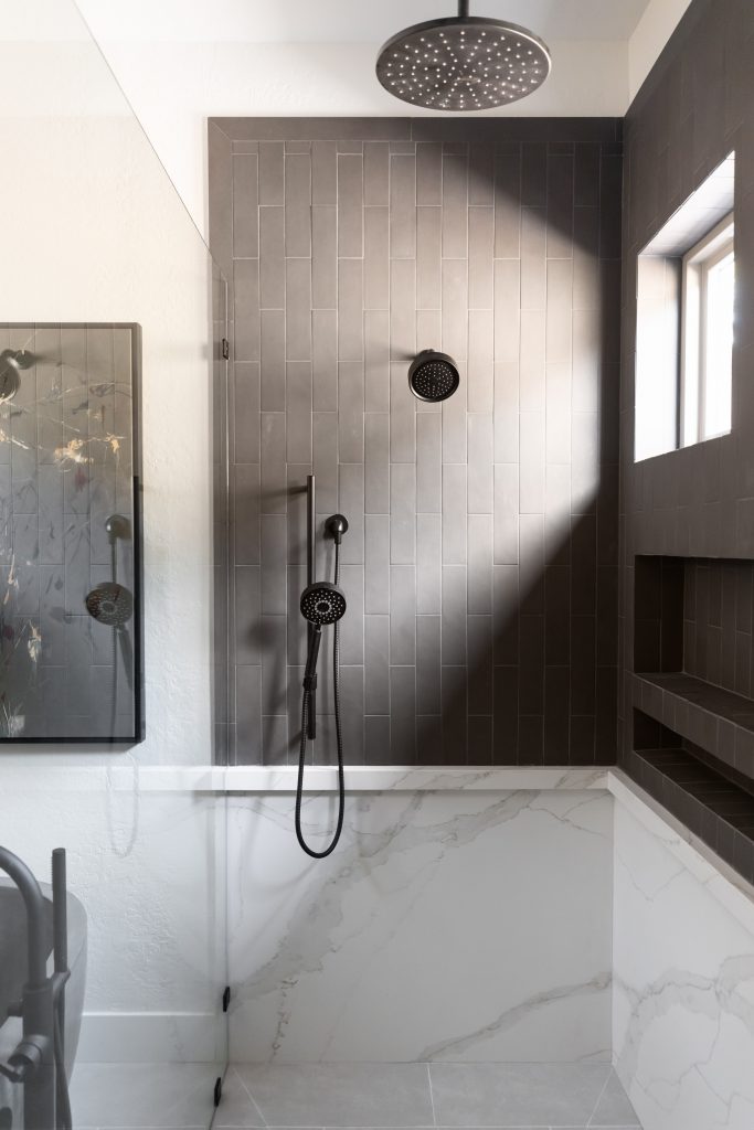 Modern shower with vertical dark tiles, black rain showerhead, handheld attachment, built-in shelf below a window, and white marble lower walls—Living with Lolo.