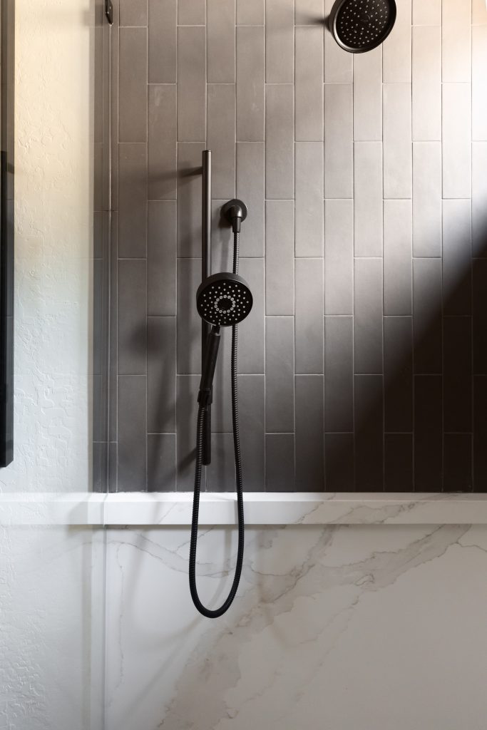 A shower with a black handheld and mounted showerhead is set against dark vertical tiles and a white marble wall. — Living with Lolo.