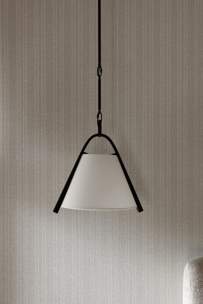 A pendant lamp with a conical white shade and black frame hangs in front of a textured beige striped wall. — Living with Lolo.