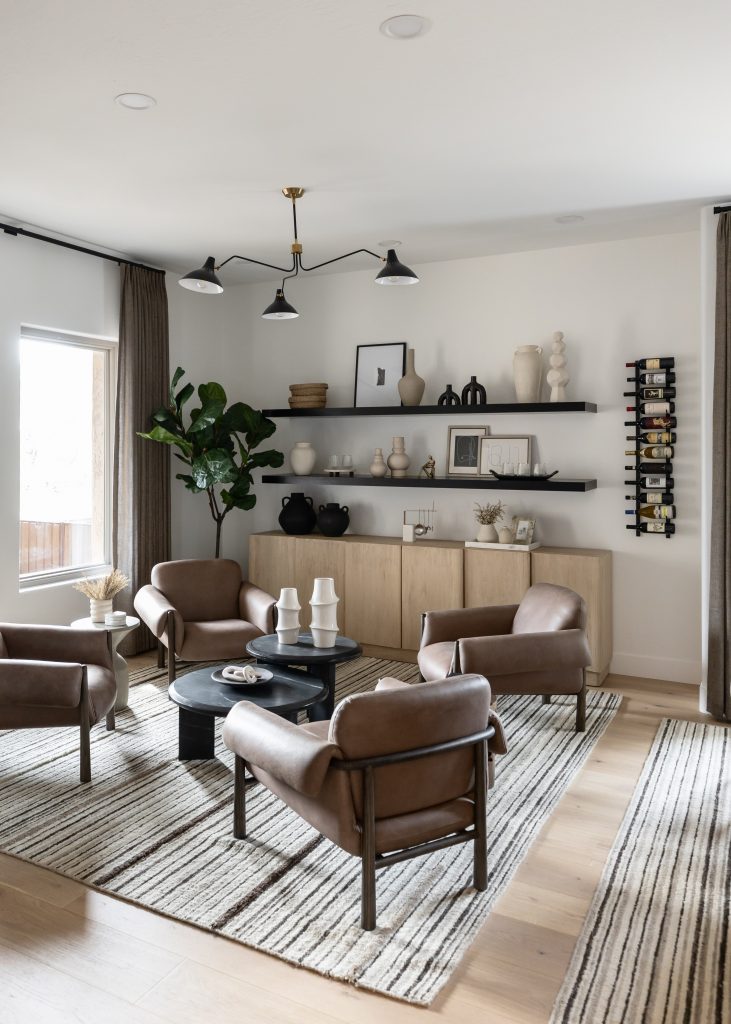 The living room has four brown leather chairs, a black round coffee table, striped rugs, wall shelves, and a floor plant by the window.— Living with Lolo.