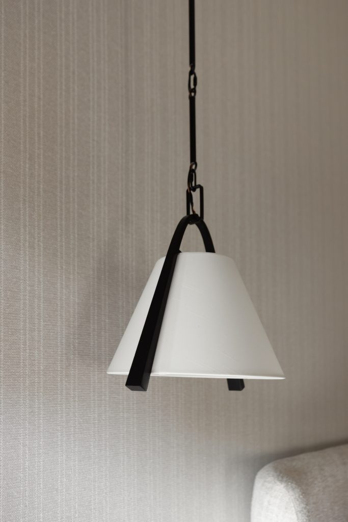 A white pendant lamp with a conical shade and black metal accents hangs above a light-colored sofa and textured beige wall. — Living with Lolo.