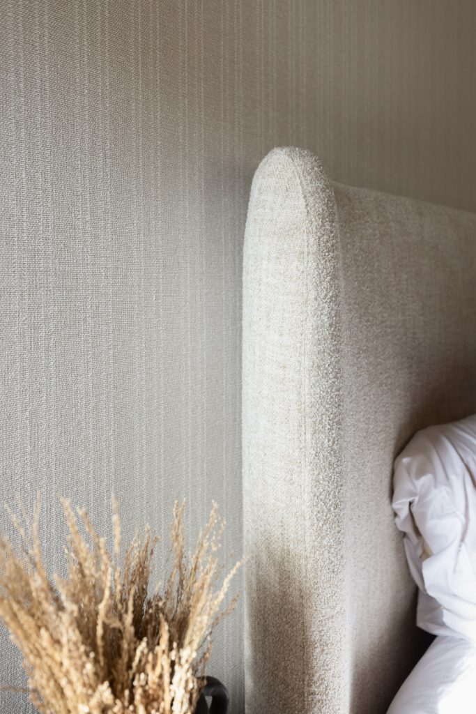 A beige upholstered headboard sits beside white bedding, with dried grass in a vase and a textured wall in the background— Living with Lolo.