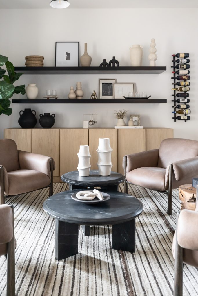 Modern living room with four brown armchairs, two black round coffee tables, a striped rug, shelves with decor, and a wine rack—Living with Lolo.