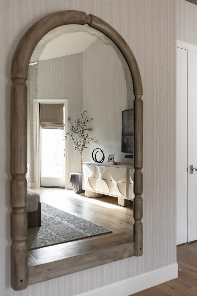 An arched wooden mirror reflects hardwood floors, a patterned sideboard, a decorative vase, a TV, and a plant near a door—Living with Lolo.