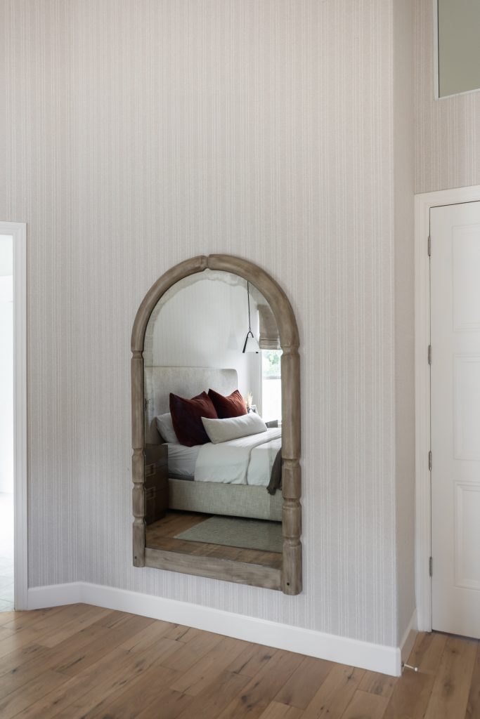 An arched mirror on a light wall reflects a bedroom with a bed, brown and white pillows, and a reading lamp—Living with Lolo.