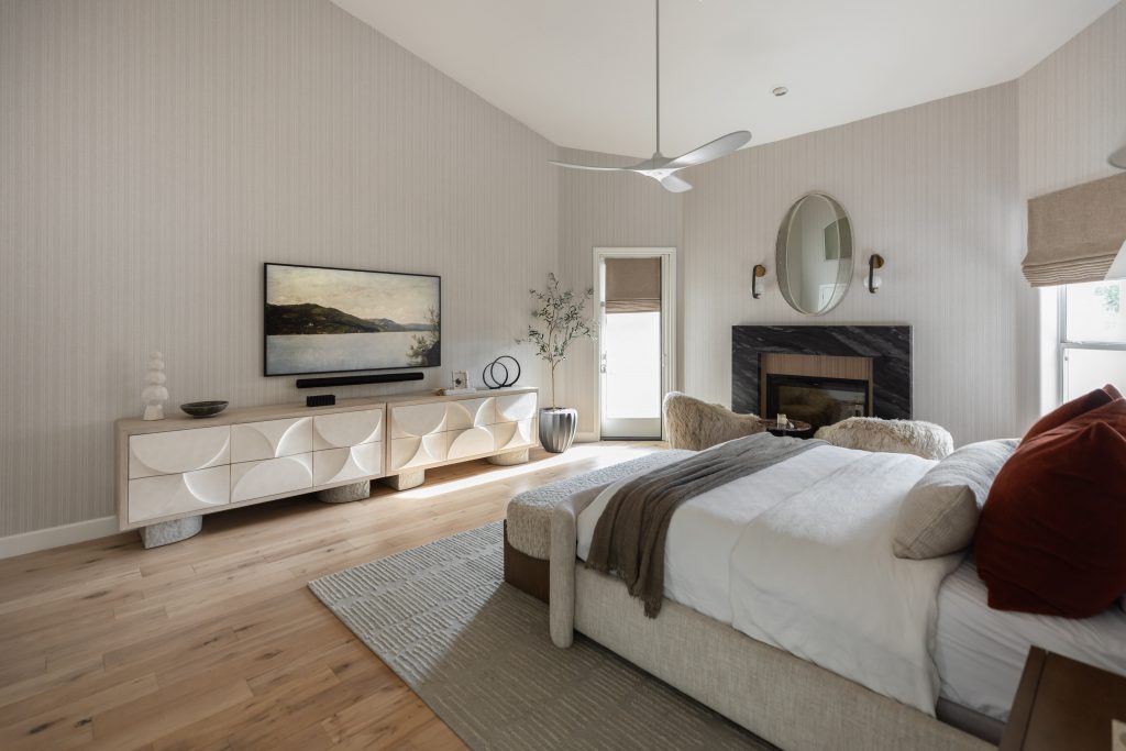 Modern bedroom featuring a bed with neutral bedding, wall-mounted TV, dresser, fireplace with round mirror, and large windows—Living with Lolo.