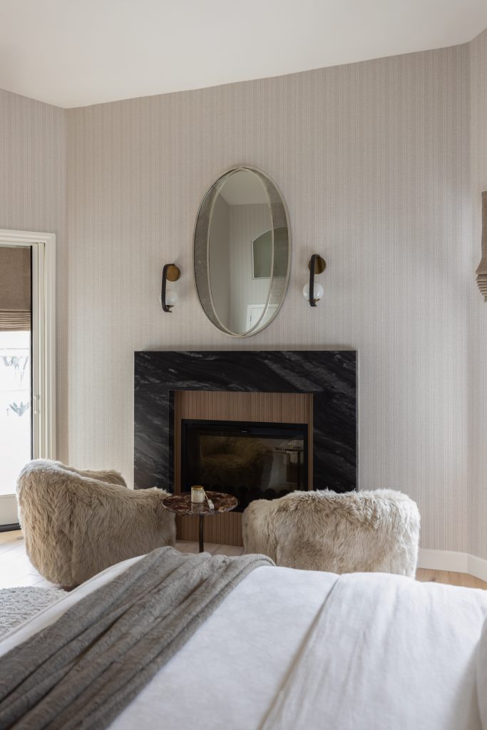 A black marble fireplace, two fuzzy armchairs, a small round table, an oval mirror, and two wall sconces are in the bedroom. — Living with Lolo.
