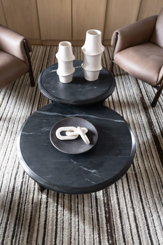 Two round black marble coffee tables with white ceramic vases and a sculpture sit on a striped rug between brown leather armchairs—Living with Lolo.