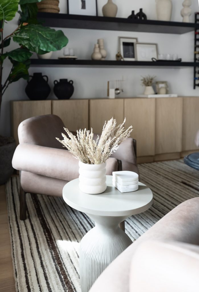 A modern living room with neutral colors features a round side table, white vase of dried flowers, two armchairs, shelves, and a striped rug — Living with Lolo.